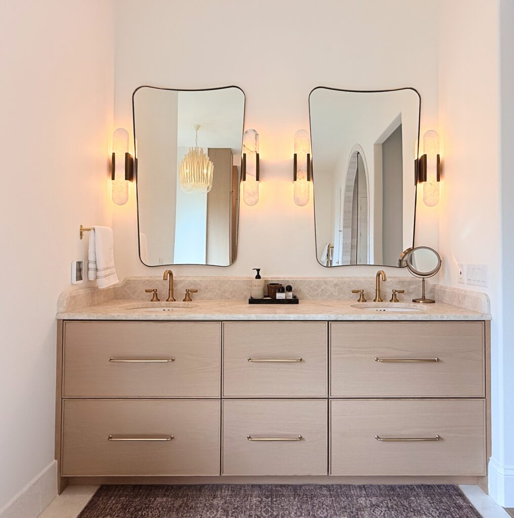 Six-drawer white oak double vanity with brass pulls, cream stone countertop, two undermount sinks and arched mirrors