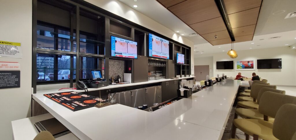 Commercial hotel bar with white quartz countertop, dark wood back bar cabinetry, glass racks and wall-mounted TVs