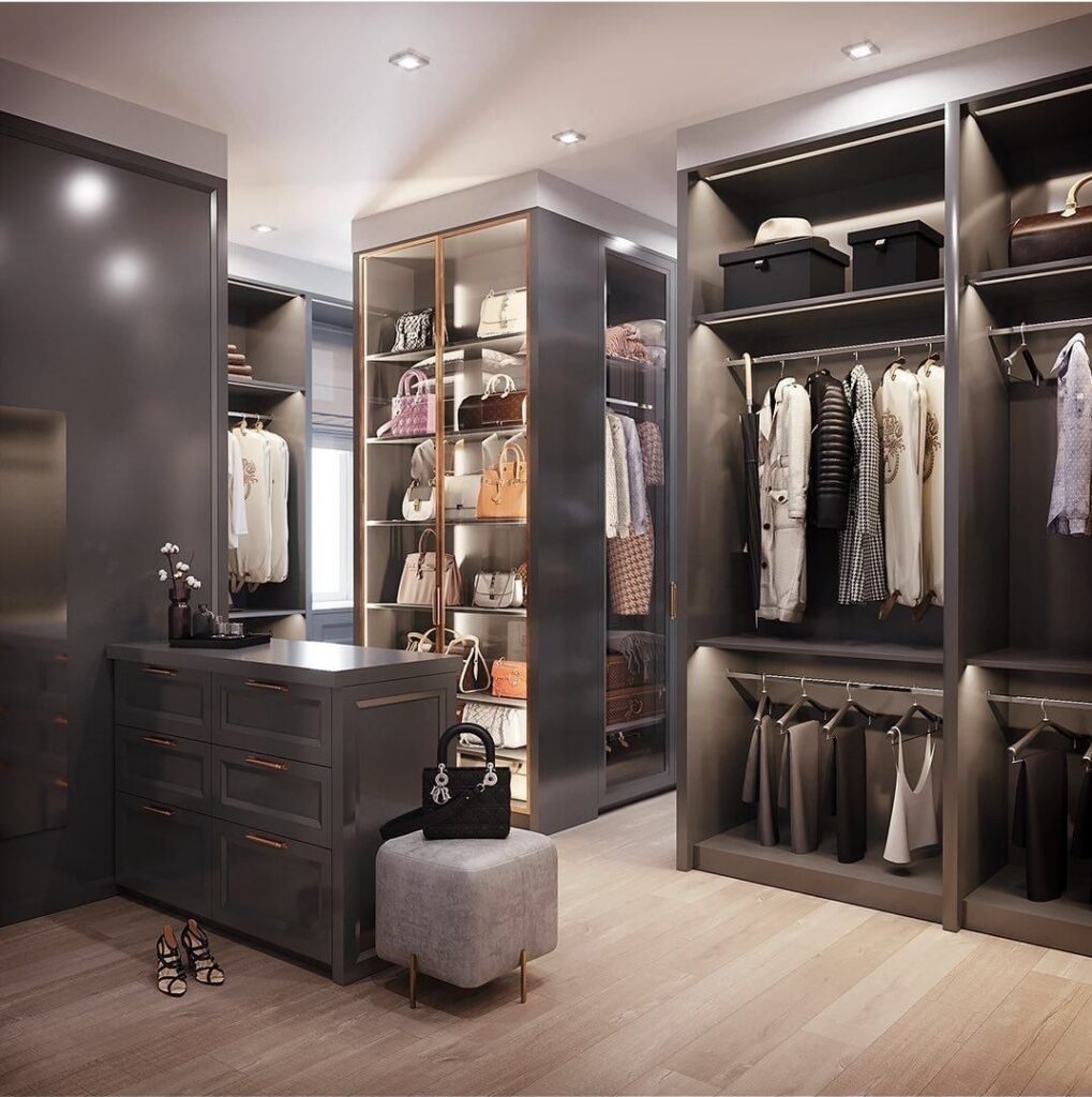 Dark gray walk-in closet with lit shelving, glass handbag cabinet, island drawers, and hanging rails