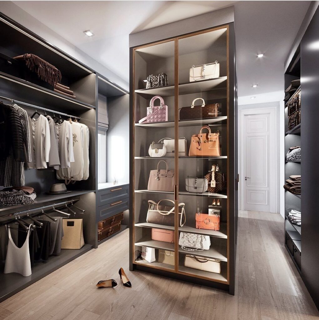 Walk-in closet with brass-framed glass handbag display cabinet, open hanging rails and lit shelving in charcoal grey