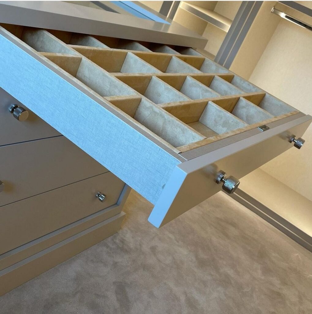 Open closet drawer with felt-lined grid dividers for watches and cufflinks, taupe fronts and chrome knobs