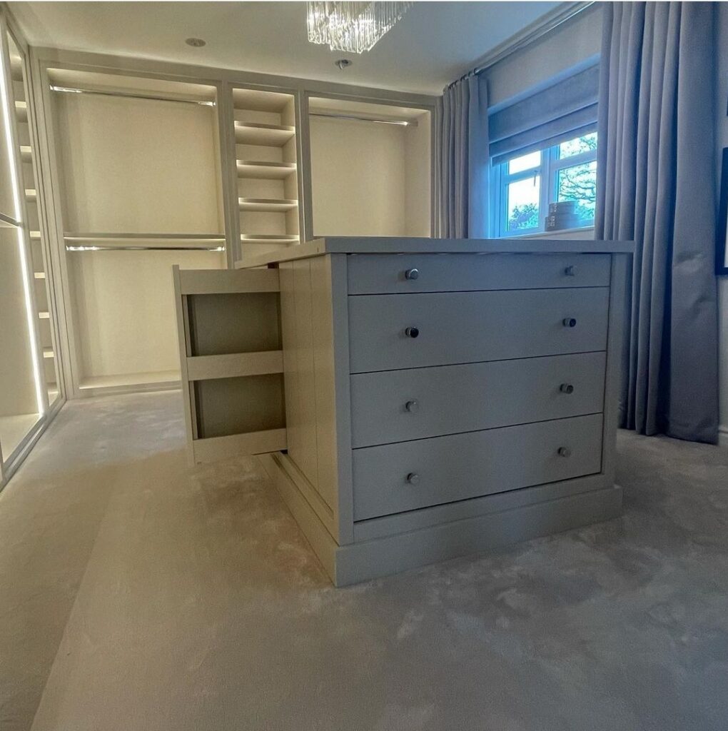 Cream walk-in wardrobe with LED-lit shelving, hanging rails and a central drawer island with open drawer
