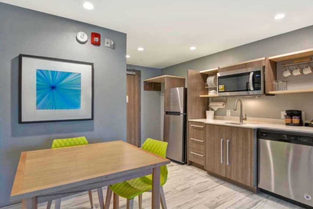 Extended-stay suite kitchenette with woodgrain slab cabinets, open shelving, stainless appliances and green dining chairs