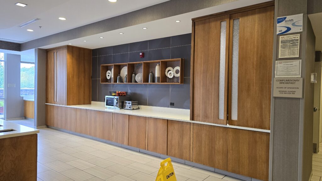 Long wood-grain hotel breakfast buffet run with white solid surface counter, open display shelving and tall doors