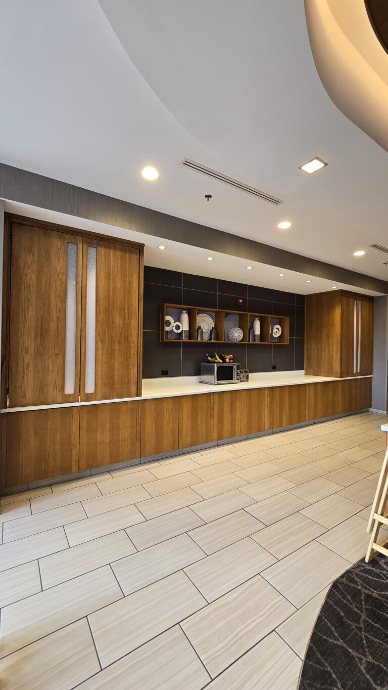 Long wood-veneer hotel buffet counter with white solid-surface top, tall pantry cabinets and lit display niches
