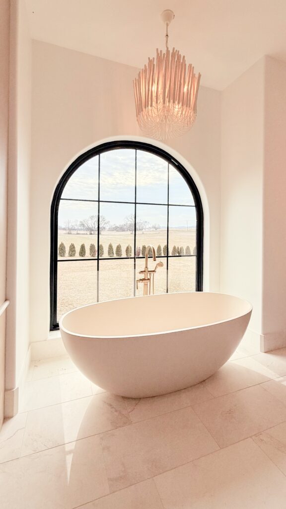 Freestanding oval soaking tub in front of a black arched grid window with brass floor filler and chandelier