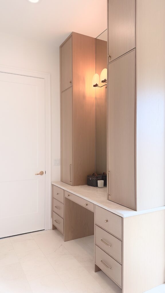 Light oak vanity with knee space, drawers, stone top and tall flanking towers beside a mirror and wall sconce